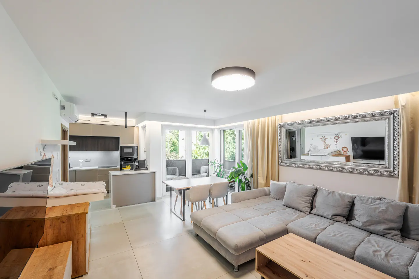 Modern open-plan living room with a grey sectional sofa, dining area, and kitchen with light-colored tile flooring.