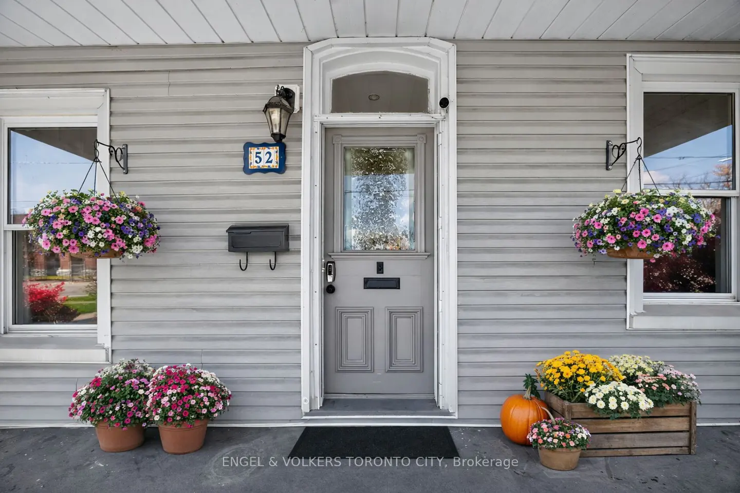 Front entrance of a grey house with number 52, hanging flower baskets, potted flowers, and a pumpkin on the porch.