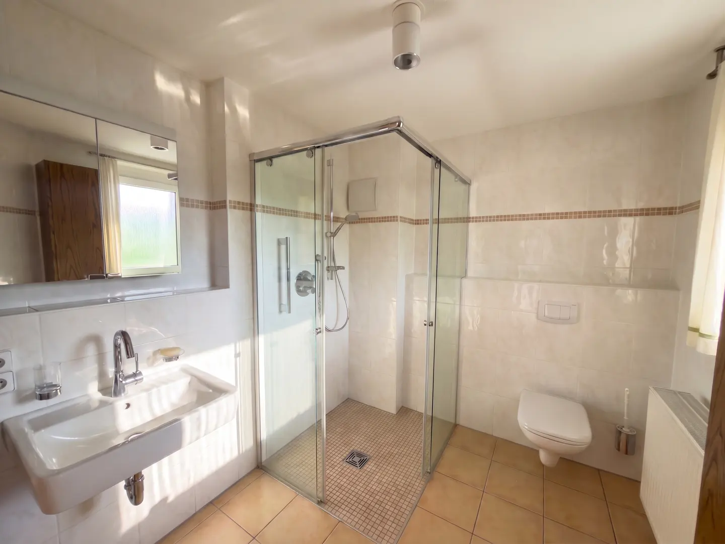 Bright bathroom with a glass shower, white sink, toilet, and beige tile floor. A mirror hangs above the sink.