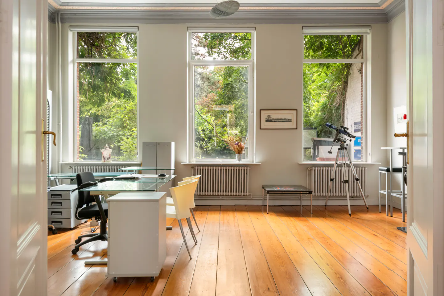 Bright office with wood floors, three large windows overlooking greenery, a glass desk, and a telescope by the window.