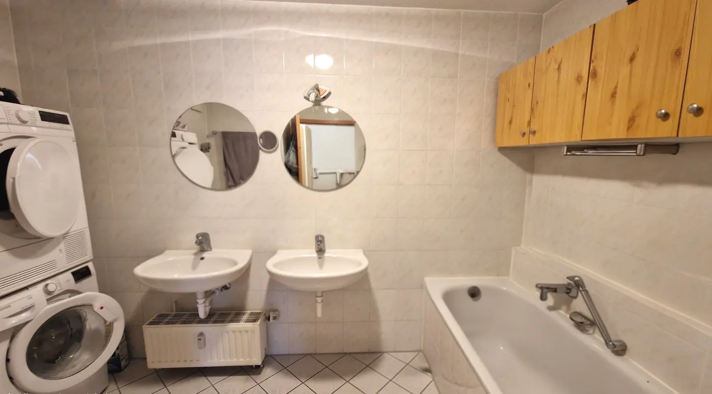Bathroom with white tile walls, two sinks, a bathtub, and a stacked washer and dryer. Two round mirrors hang above the sinks.
