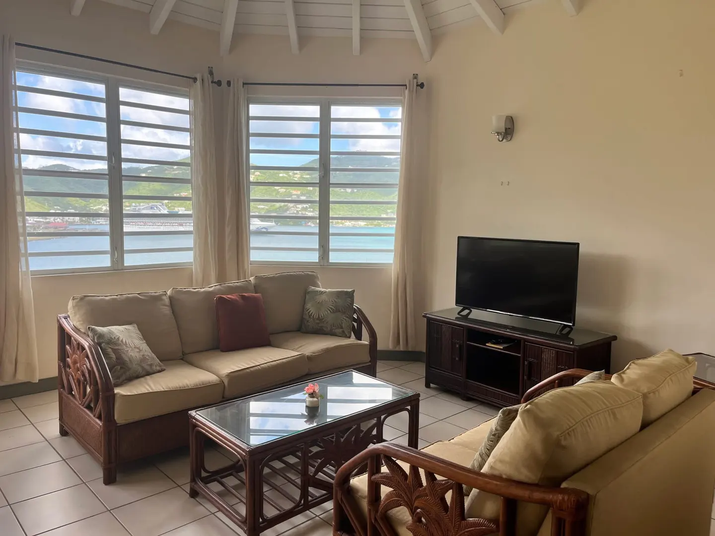 Living room with rattan furniture, beige walls, and a view of the ocean through large windows. A TV sits on a dark wood stand.