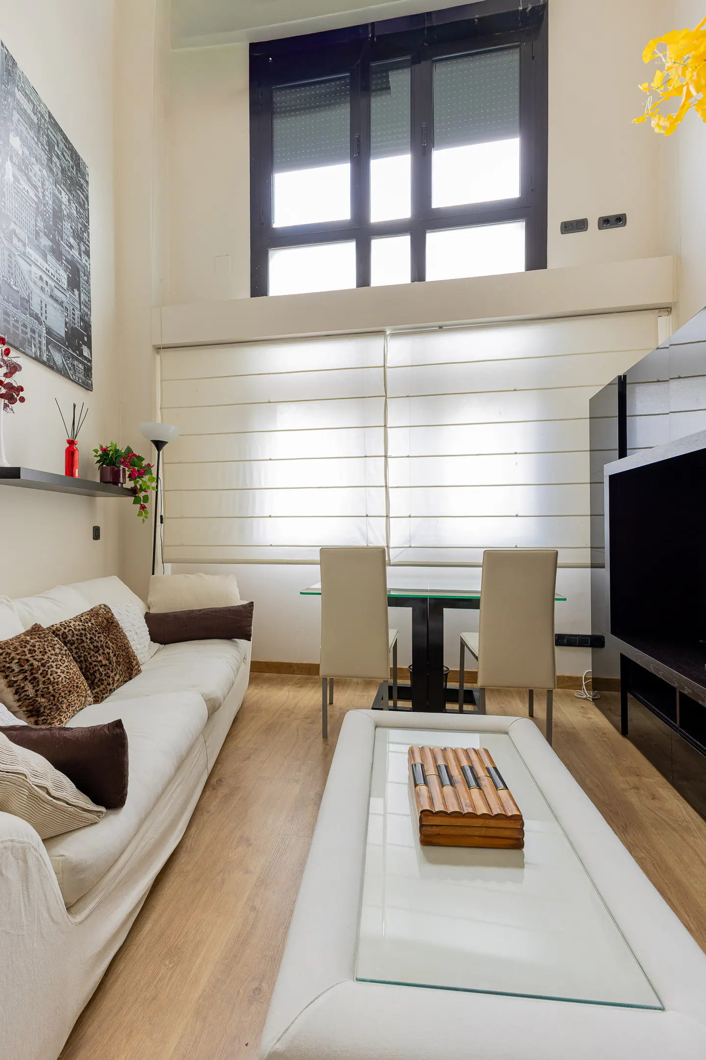 Bright living room with white sofa, glass coffee table, and dining set. Large window with blinds, and modern art on the wall.
