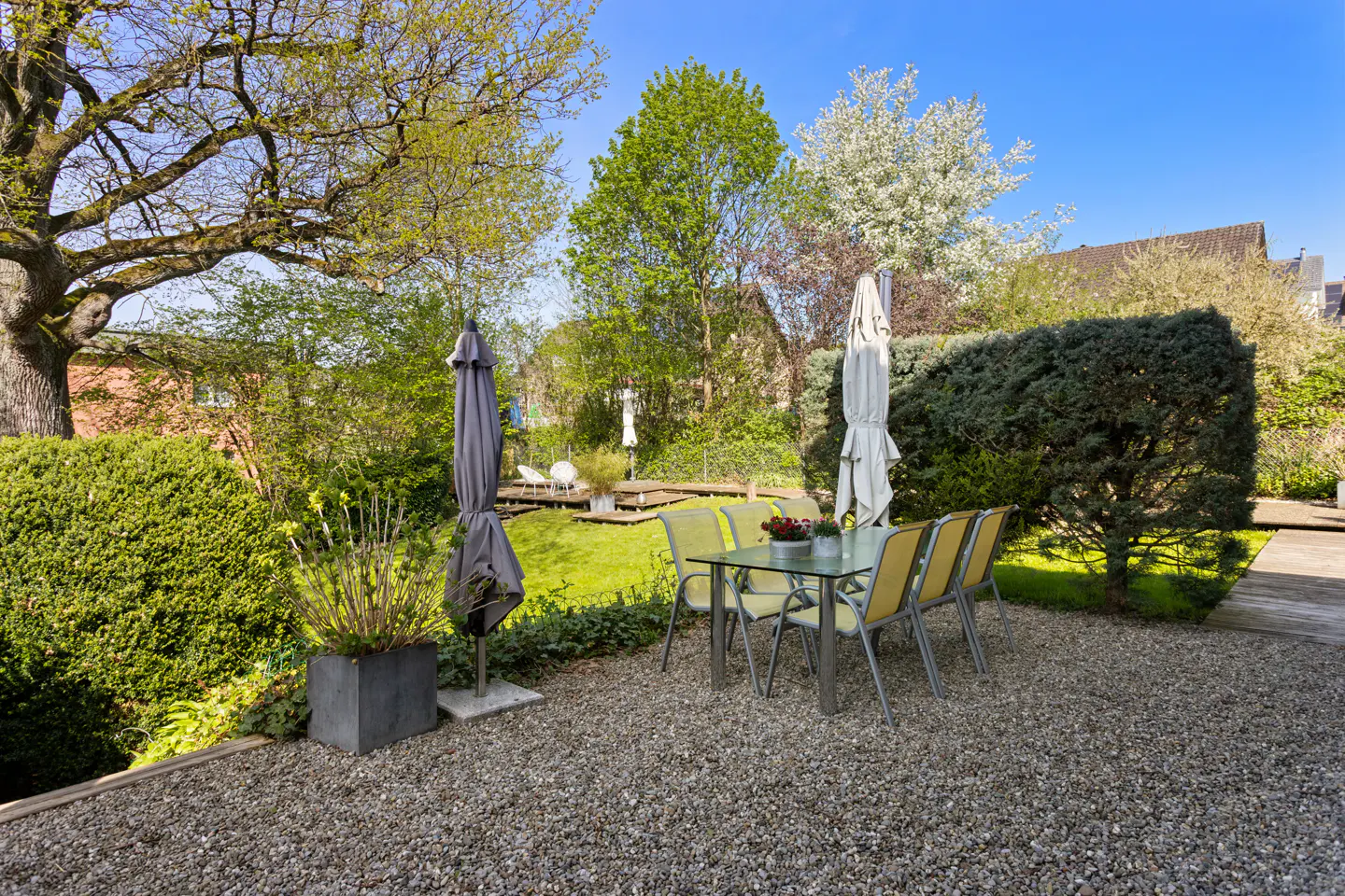 A backyard patio with a glass table and yellow chairs sits on a gravel surface, surrounded by green bushes and trees.