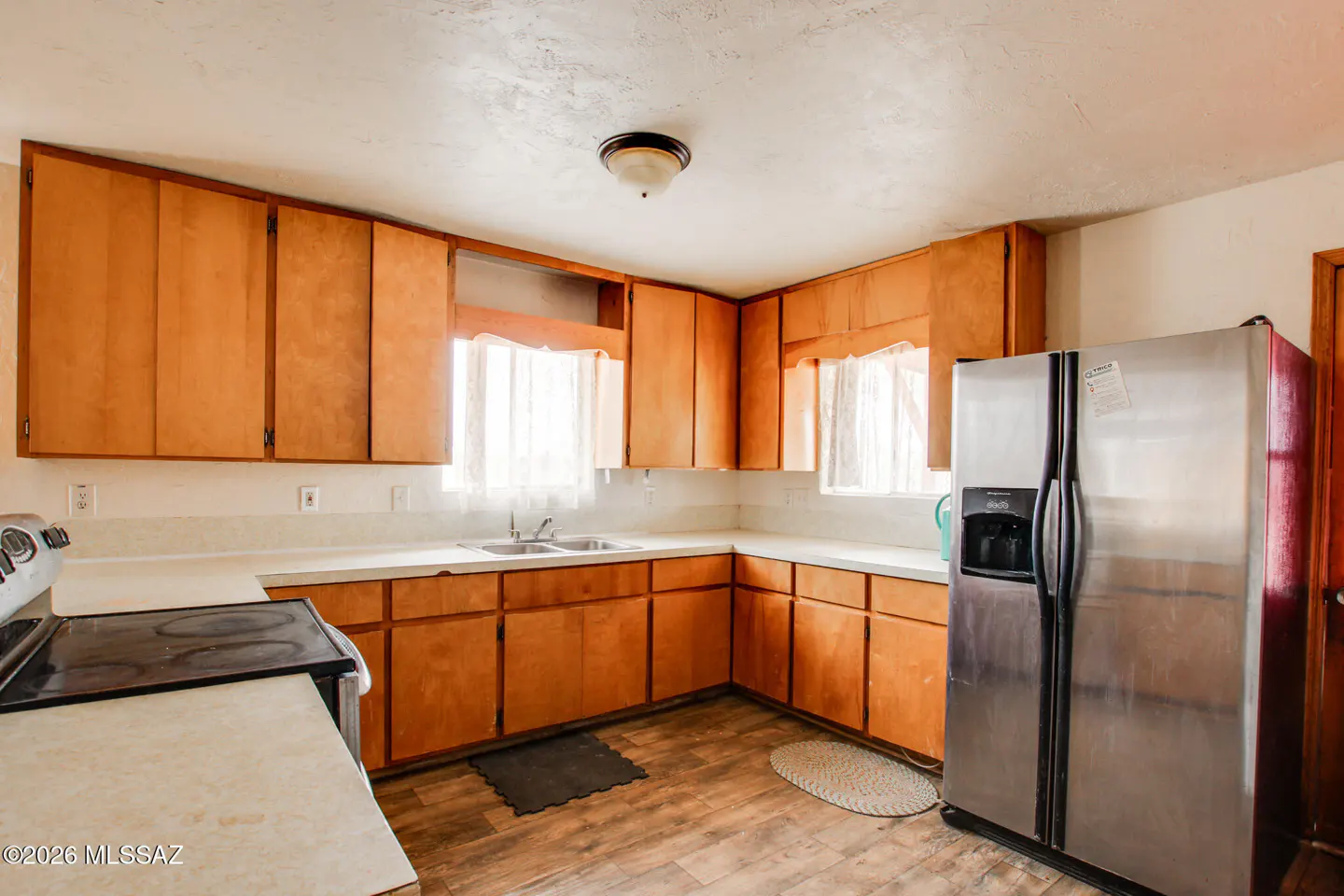 A kitchen with light wood cabinets, white countertops, and a stainless steel refrigerator. A window with sheer curtains is above the sink.