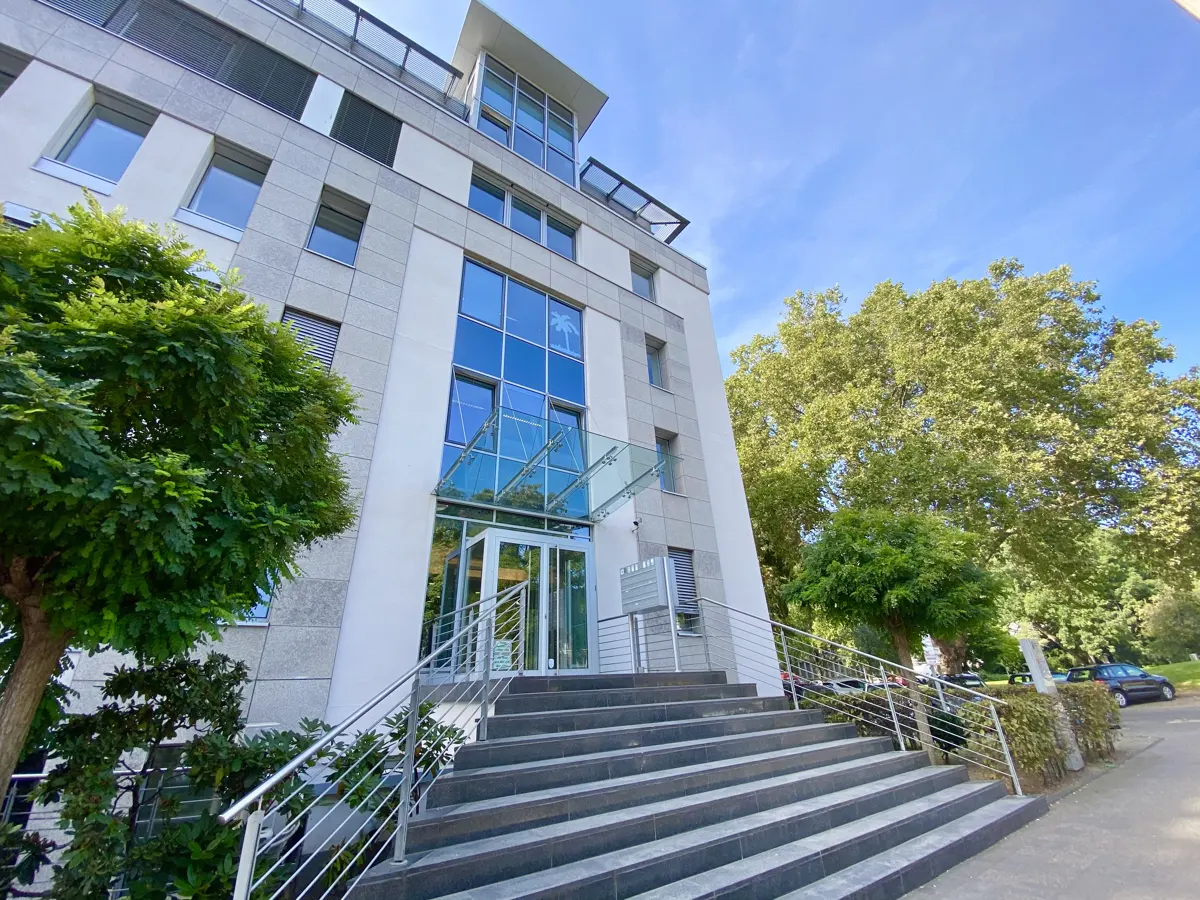 Exterior view of a modern, multi-story office building with gray stone and white accents, accessed by a wide staircase with metal railings.