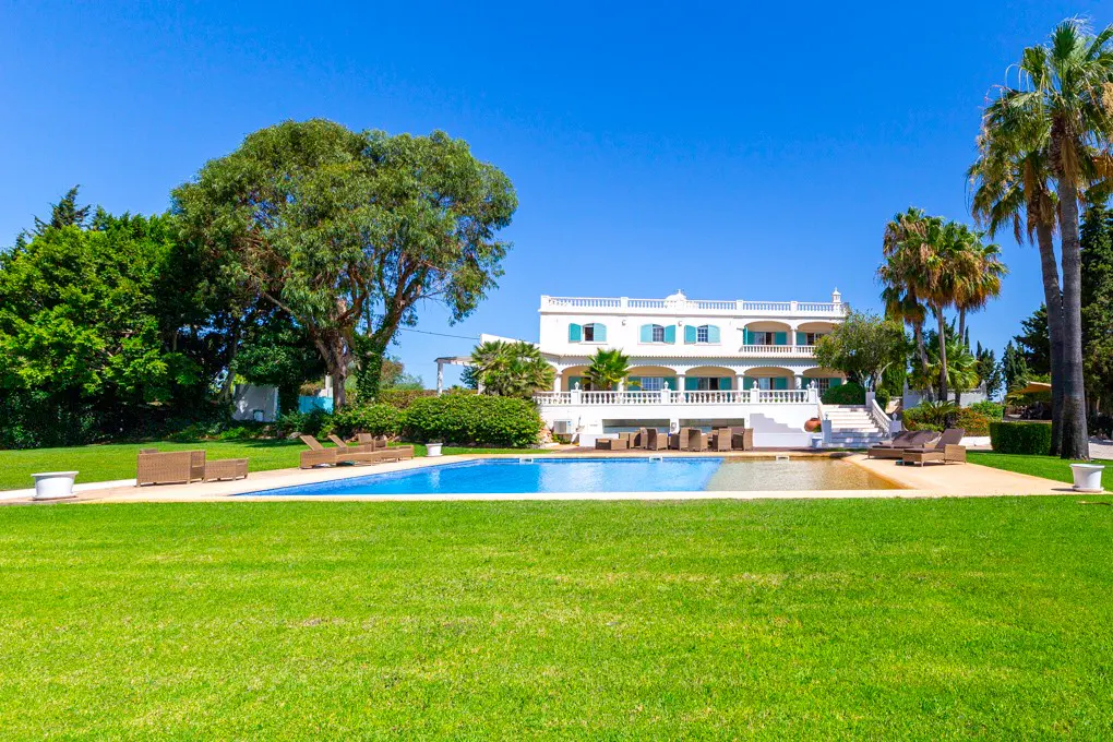 A white two-story house with a blue pool and green lawn under a clear blue sky. Palm trees and other greenery surround the property.