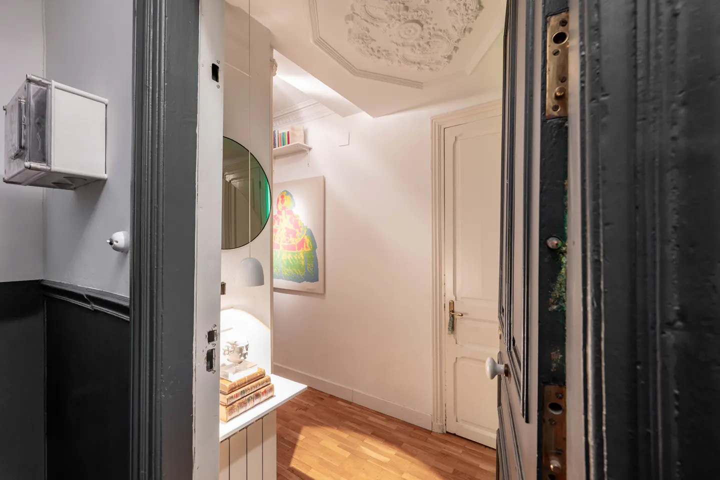 View through a dark gray door into a bright hallway with wood floors and white walls. A round mirror and colorful art hang on the wall.