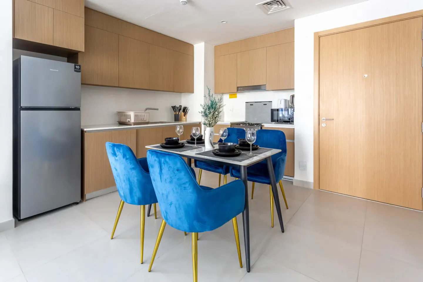 A modern kitchen with light wood cabinets, a stainless steel refrigerator, and a dining table with four blue velvet chairs.