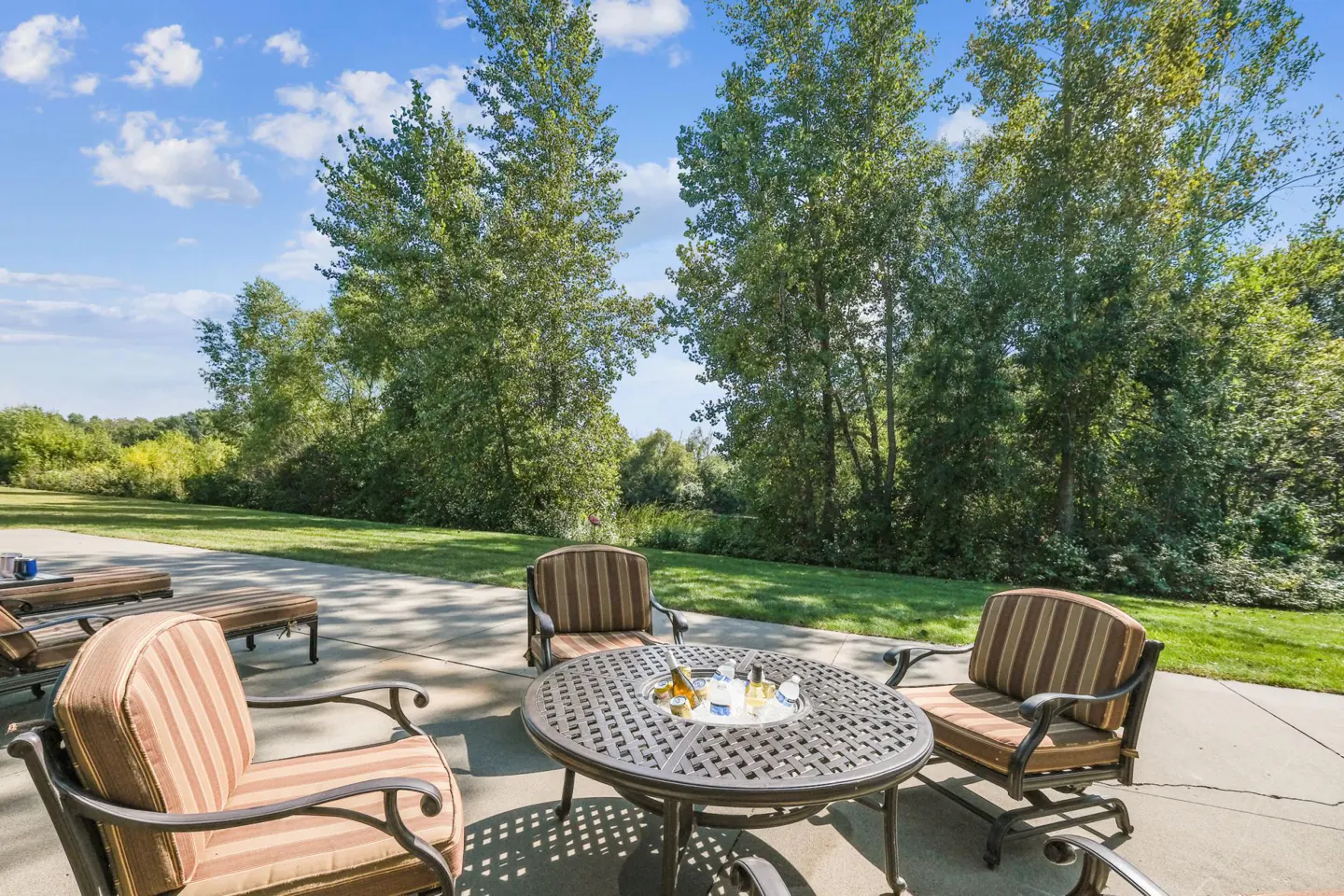 Outdoor patio with striped chairs and a metal table holding drinks on ice, set against a backdrop of green trees and a blue sky.