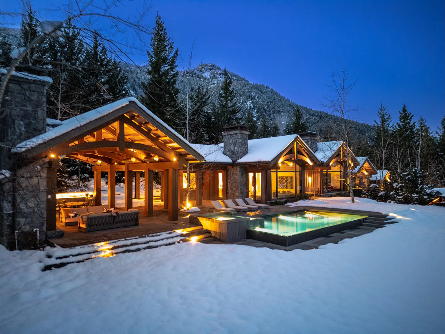 Exterior view of a luxury home with a pool and outdoor living area in a snowy, wooded setting at dusk.
