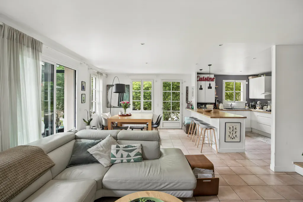 Open-concept living space with a white sectional sofa, dining table, and kitchen with a breakfast bar. Large windows and doors let in natural light.