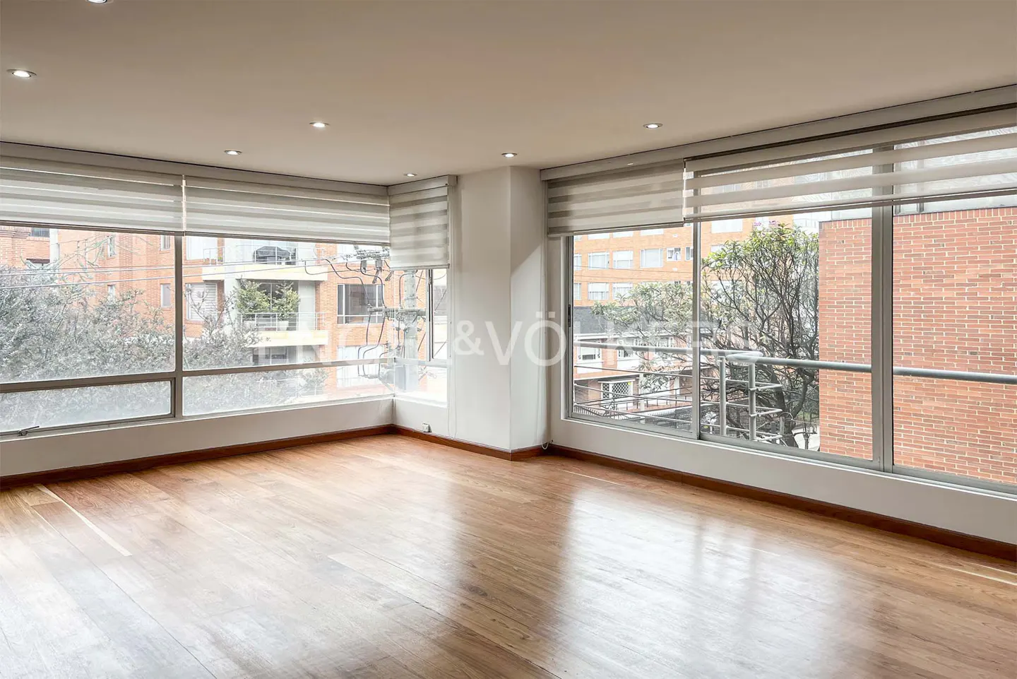 Bright, empty room with wood floors and large windows. Outside view of trees and brick buildings. Blinds are partially drawn.