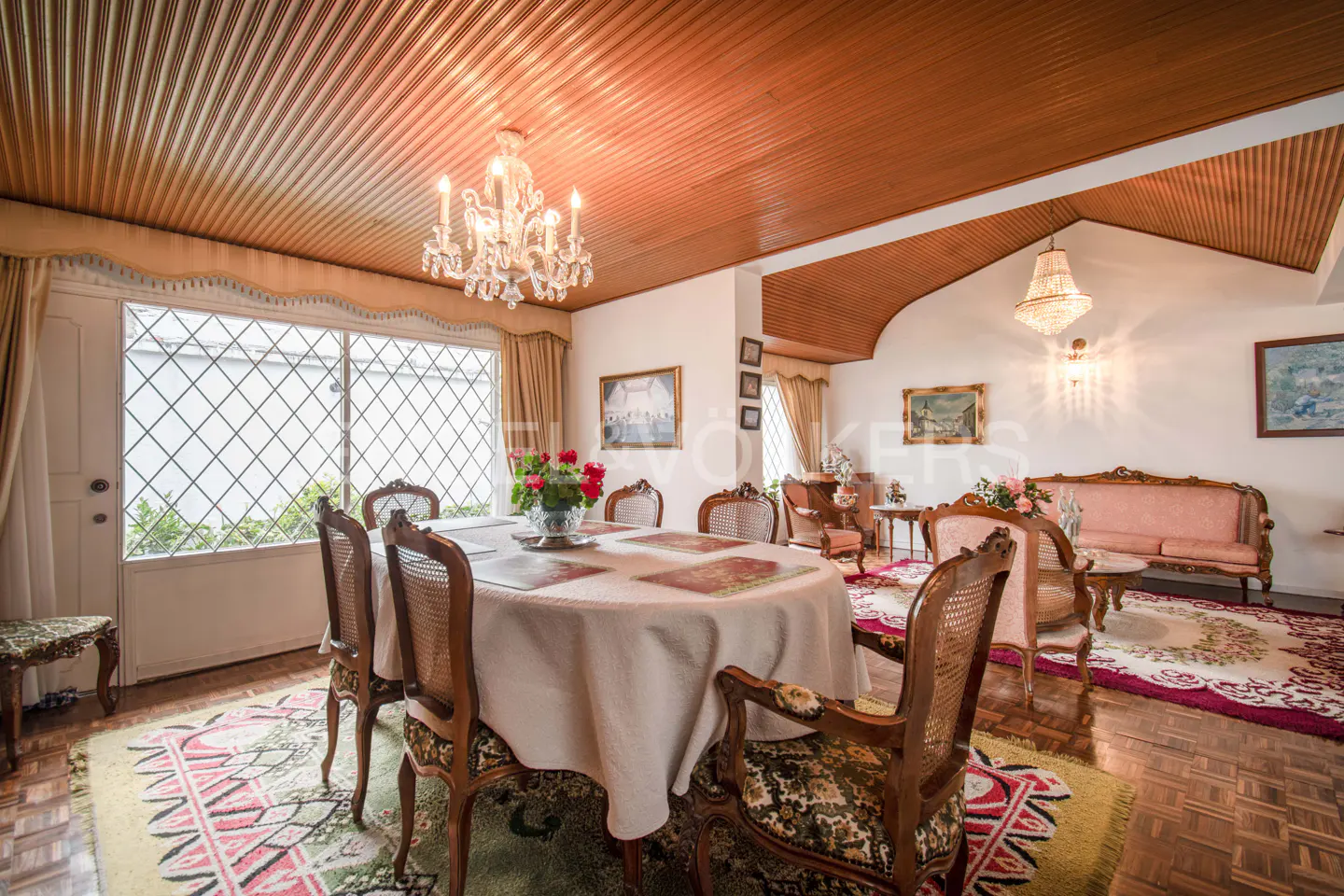 A formal dining room with a round table, chandelier, and wood-paneled ceiling. A sitting area is visible in the background.