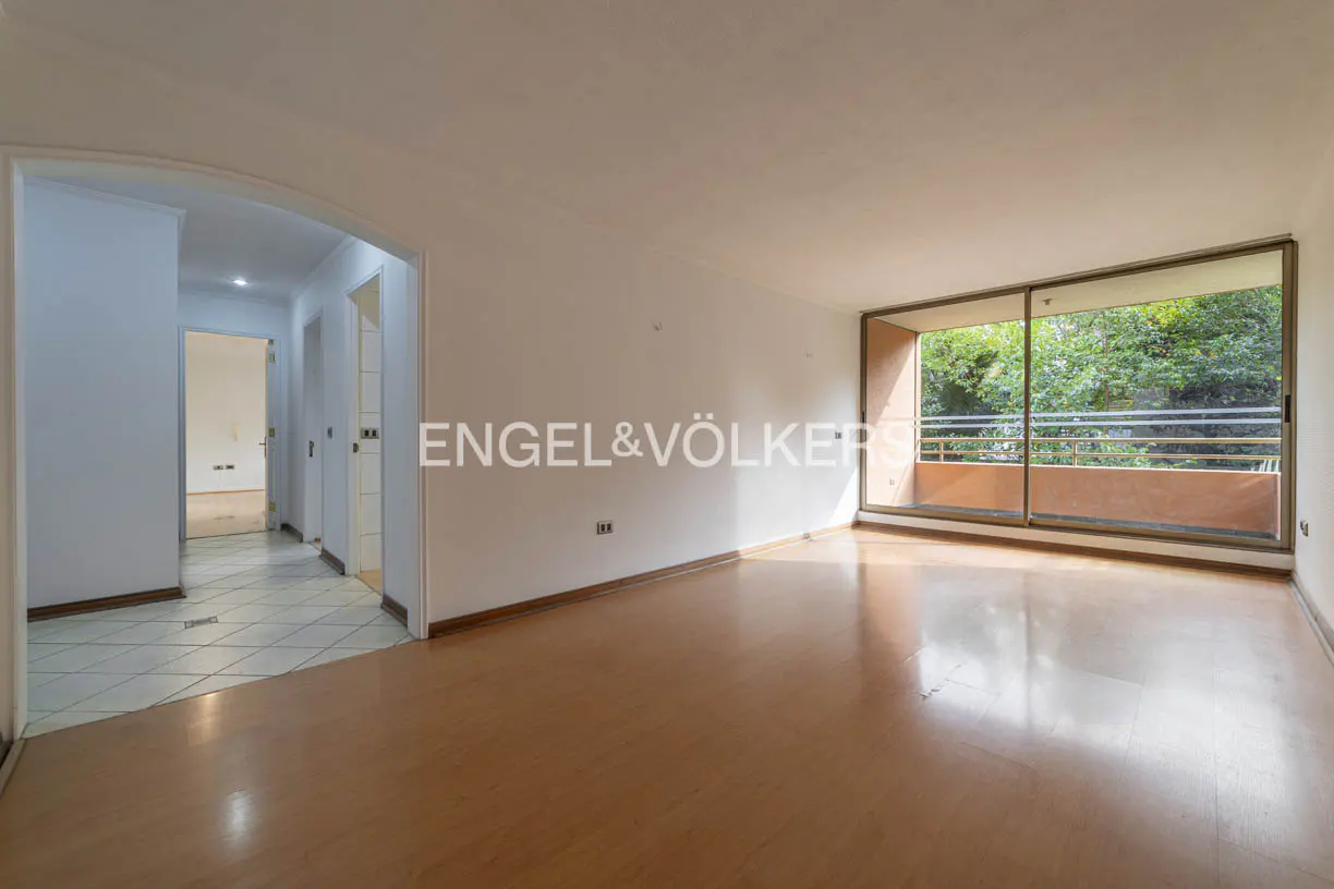 Empty living room with wood floors, white walls, and a large window overlooking trees. A hallway leads to other rooms. Engel & Völkers logo on the wall.