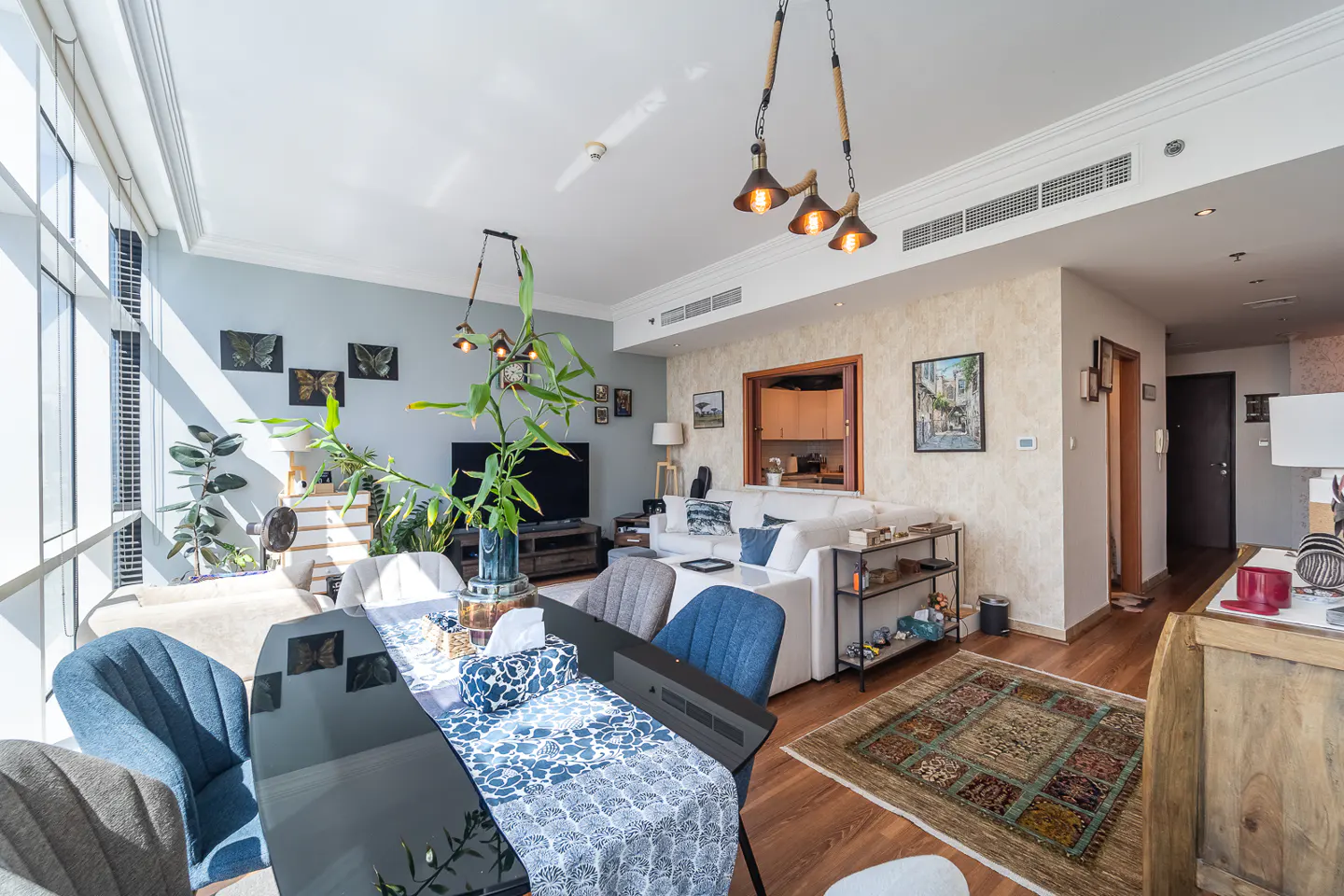 Bright, modern living room with a black dining table, blue chairs, white sofa, and wooden floors. Plants and artwork add a touch of nature.