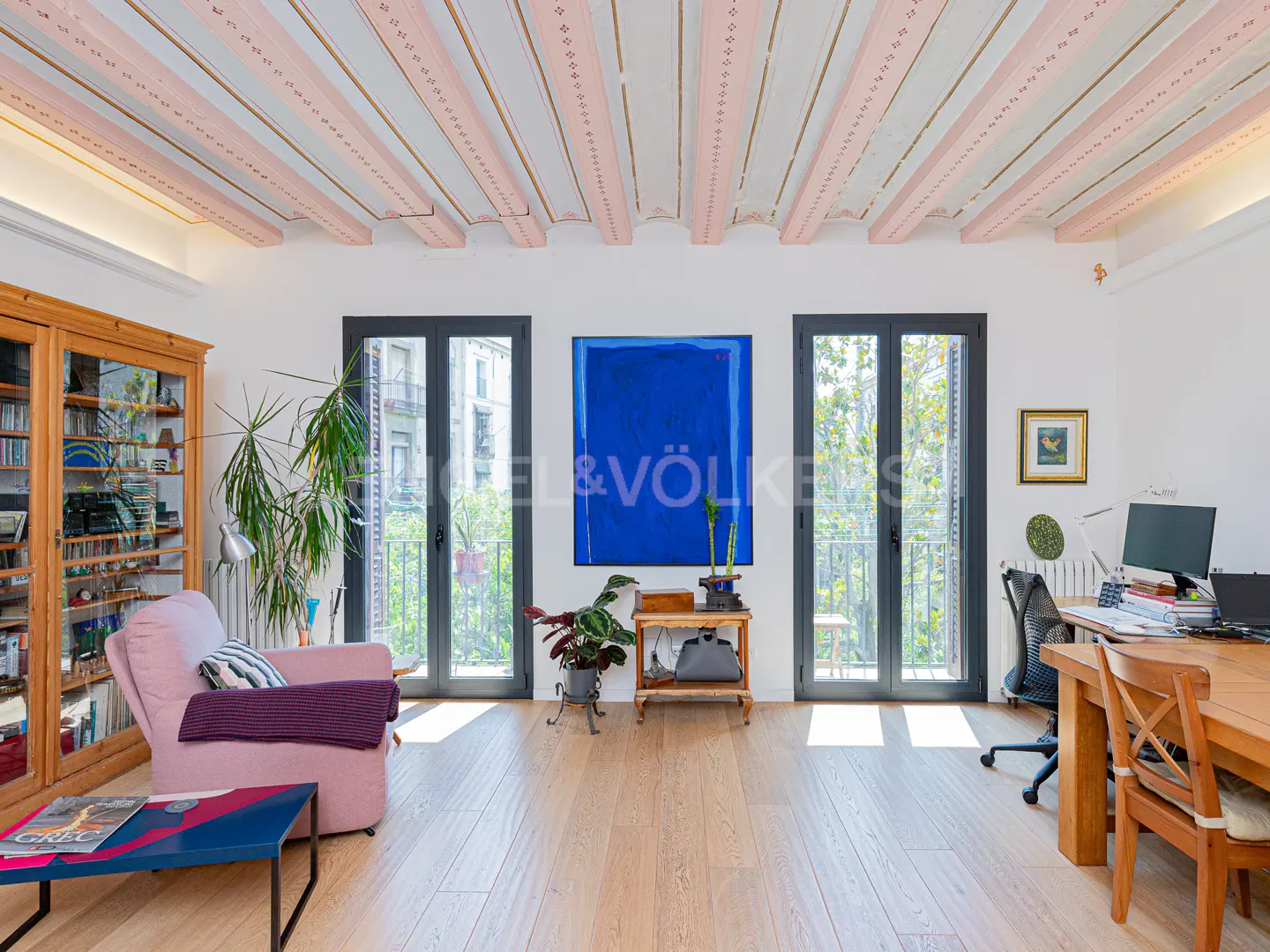 Bright living room with pink ceiling beams, wood floors, and two sets of French doors. A blue painting hangs between the doors.