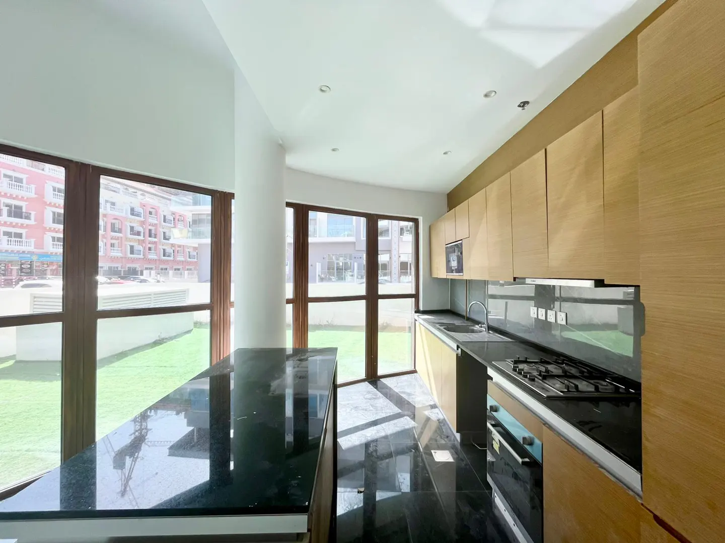A modern kitchen with light wood cabinets, black countertops, and a large island. Windows offer a view of a green lawn and buildings.