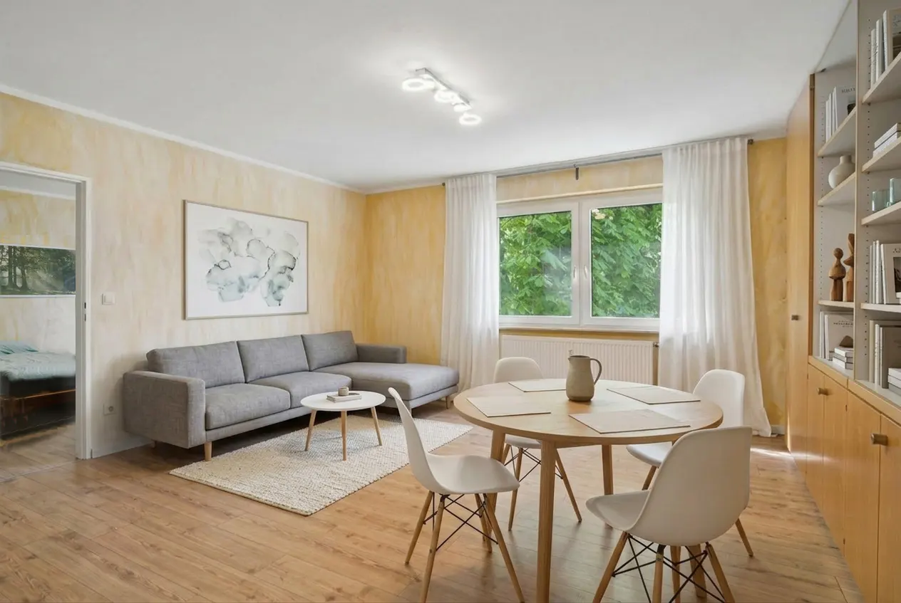 Living room with a gray sofa, round table with white chairs, and a window with white curtains. A painting hangs above the sofa.