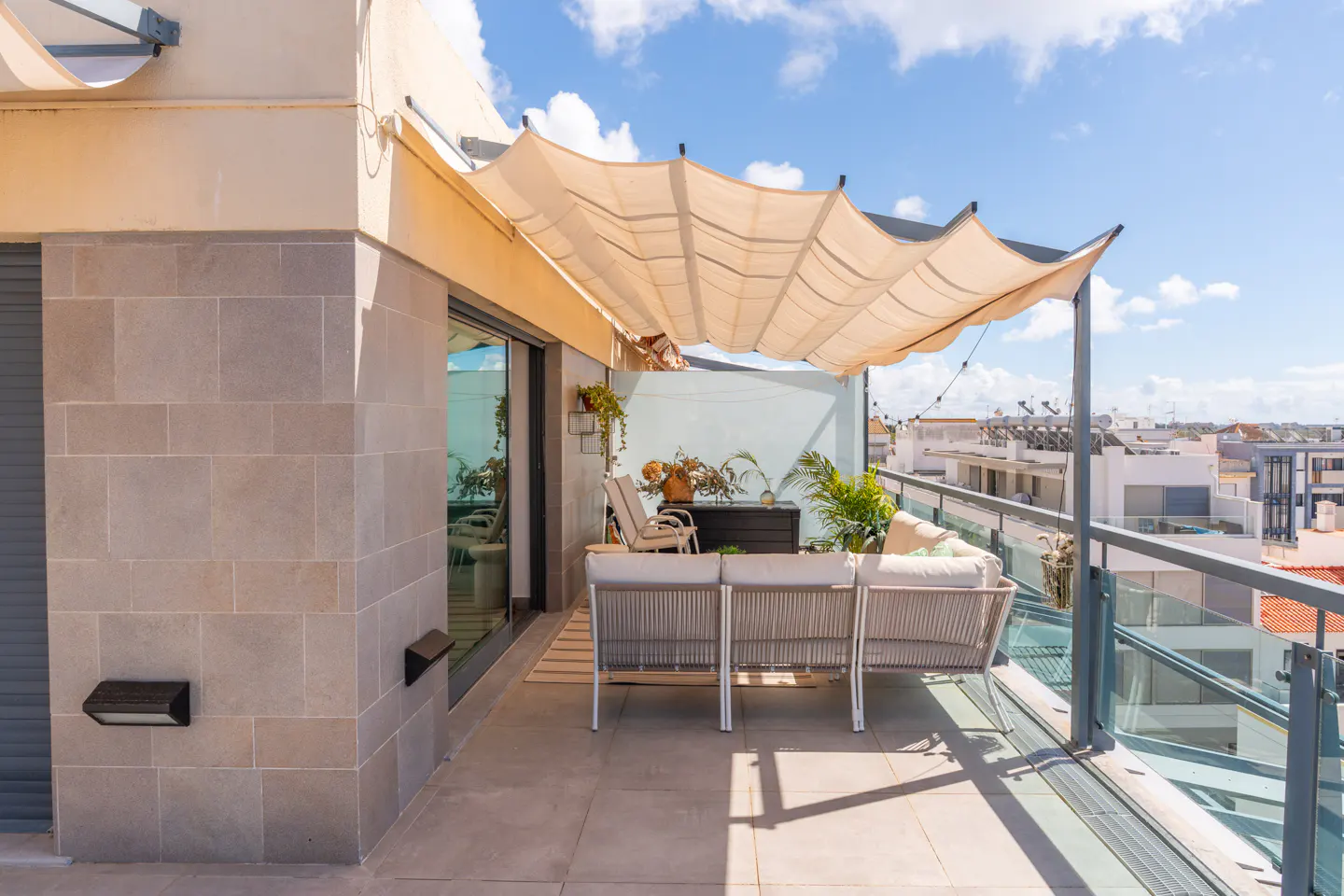A sunlit balcony with a beige awning, a gray sofa, and a glass railing overlooking a cityscape under a blue sky.