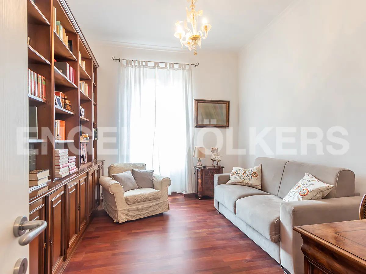 Living room with a large bookcase, a beige sofa, and an armchair on a red wood floor. A chandelier hangs from the ceiling.