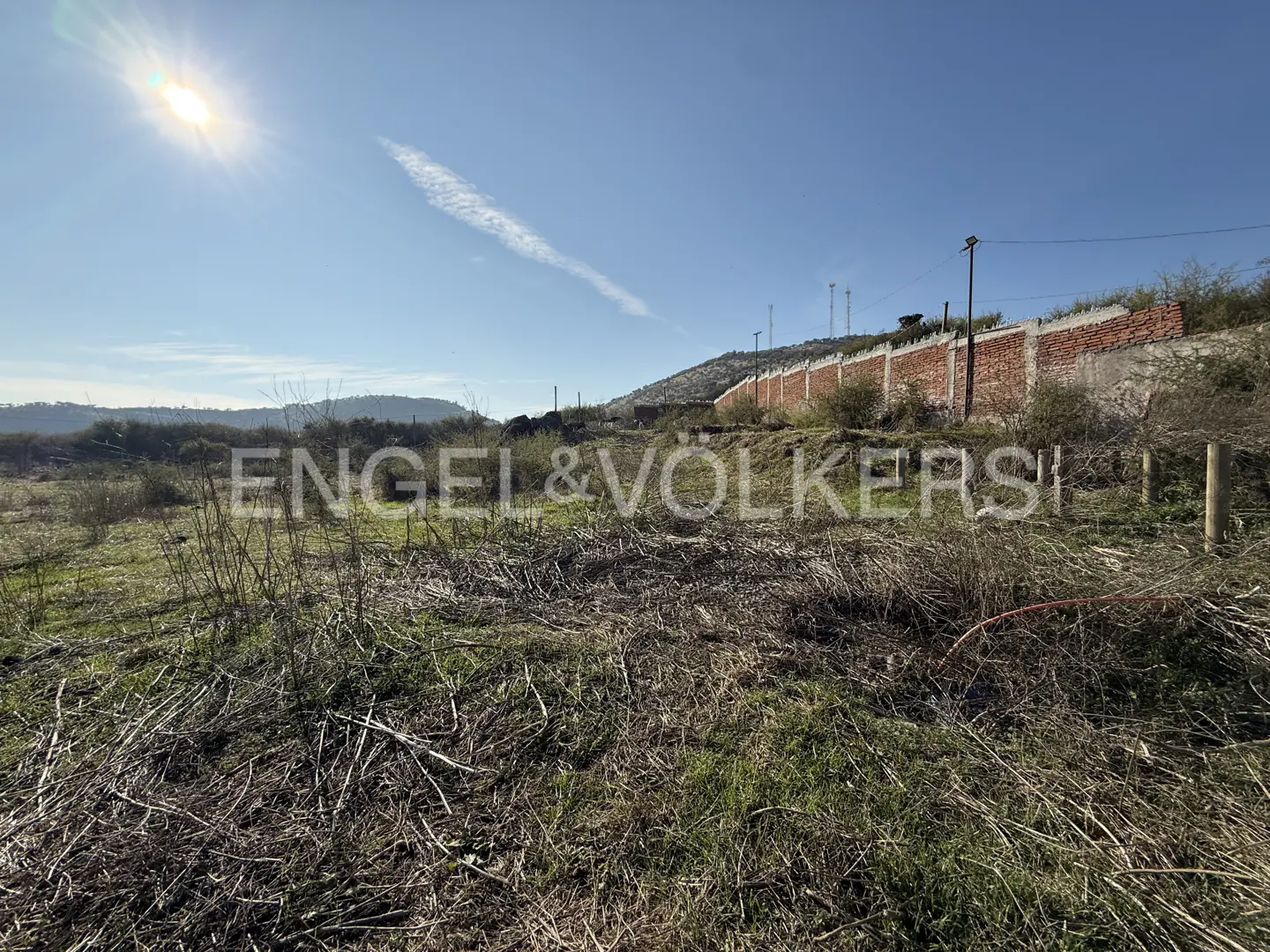 Vacant land with dry grass and a brick wall under a blue sky with the sun shining.