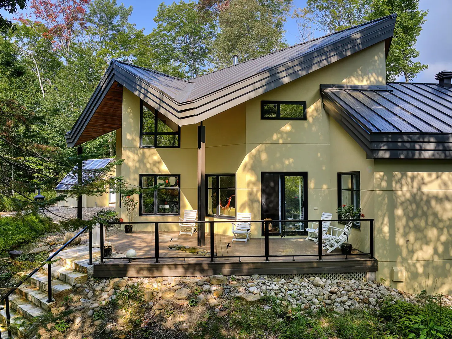 Exterior view of a two-story yellow house with a metal roof, black trim windows, and a deck with white chairs.