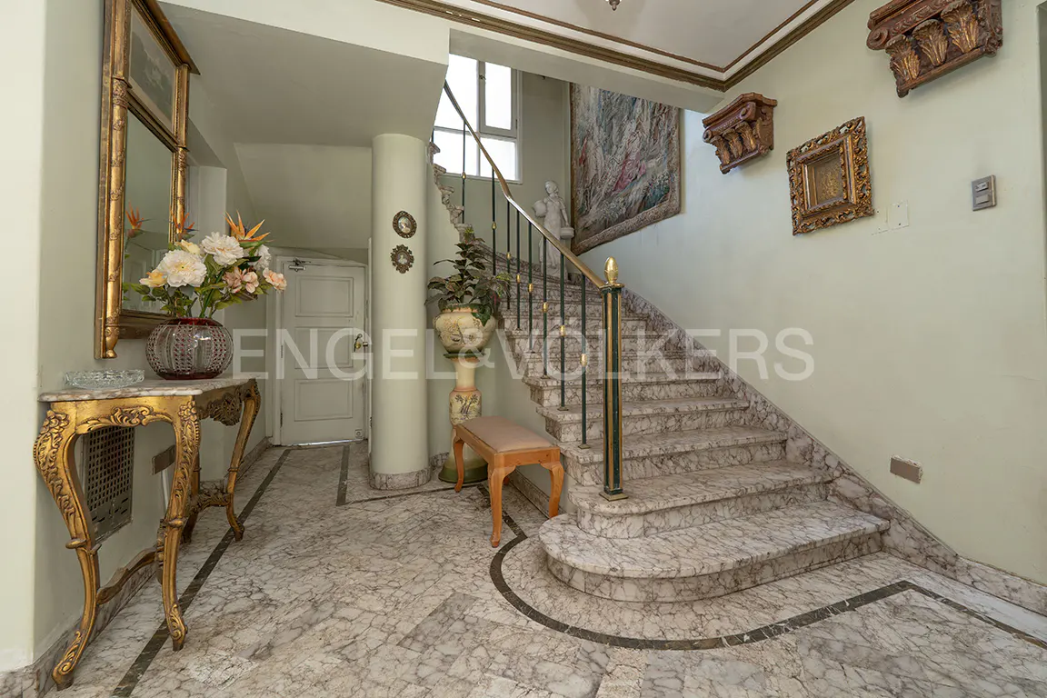 Foyer with marble floors, staircase, and gold accents. A table with flowers sits near a mirror. Tapestry hangs on the wall.