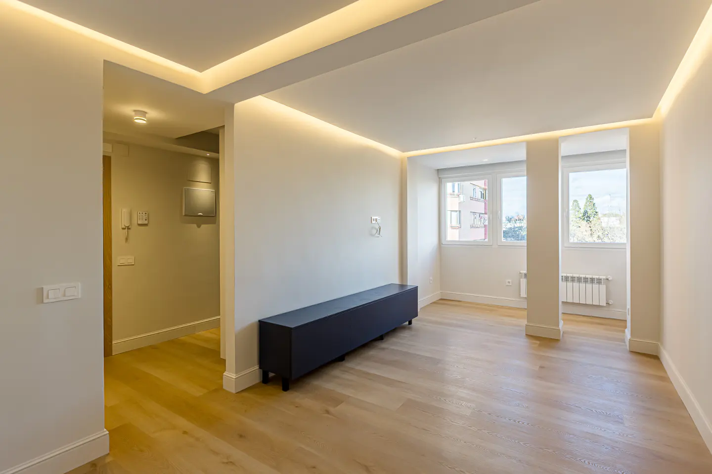 Bright, modern living room with wood floors, white walls, and recessed ceiling lights. A dark blue cabinet sits against the wall. Windows offer a view of a building and trees.