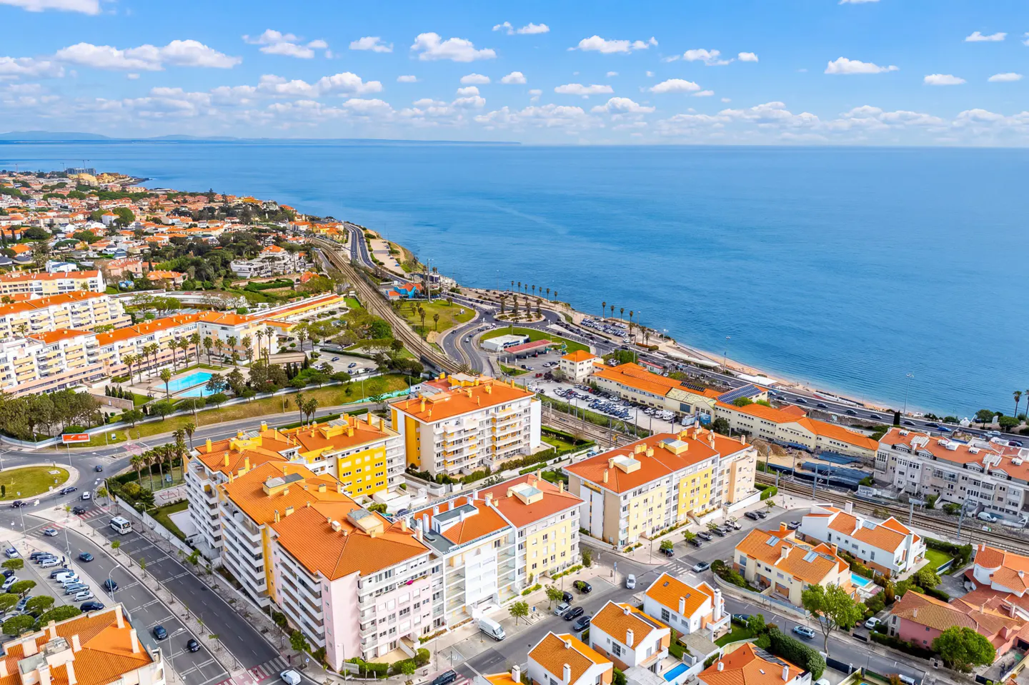 Aerial view of a coastal town with orange-roofed buildings, a railway, and the blue ocean under a partly cloudy sky.