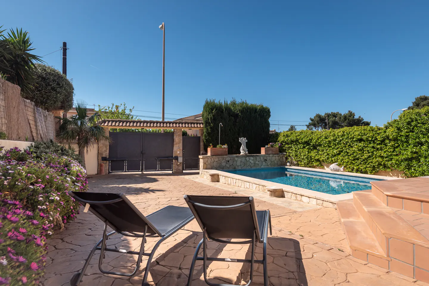 Two black lounge chairs on a stone patio beside a rectangular swimming pool, surrounded by lush greenery and a metal gate.