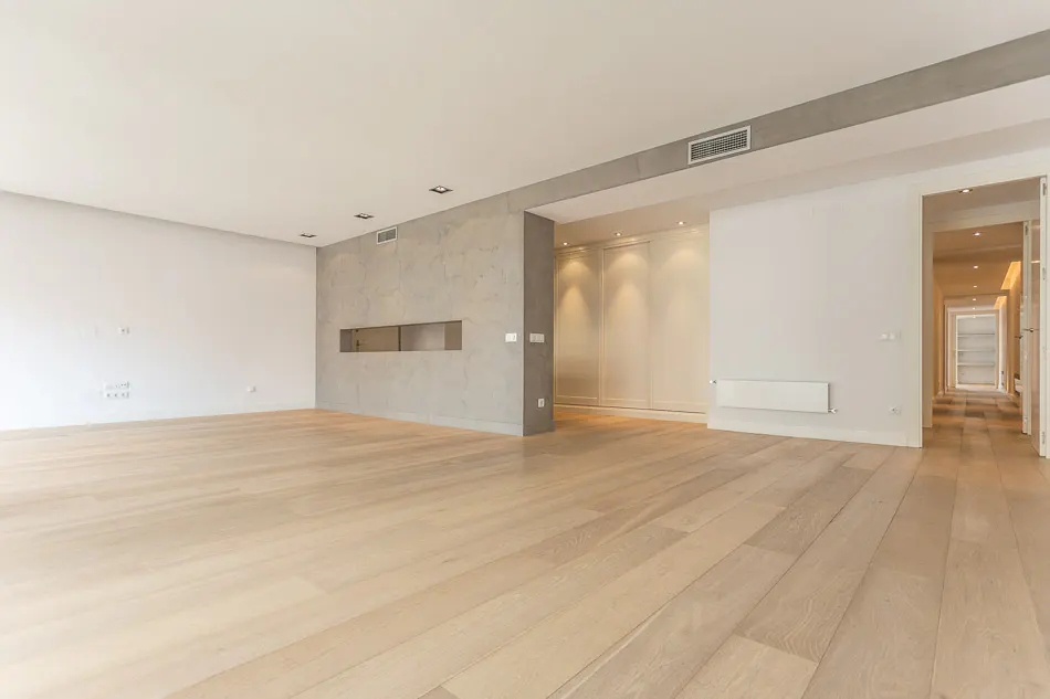 Bright, empty room with light wood floors, white walls, and a gray accent wall with a rectangular niche. A hallway is visible in the background.