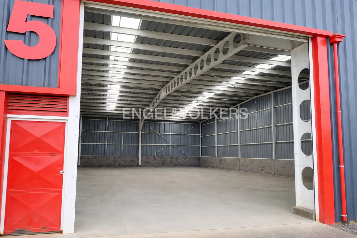 Empty industrial warehouse with grey metal walls, concrete floor, and a large red door marked with the number 5.