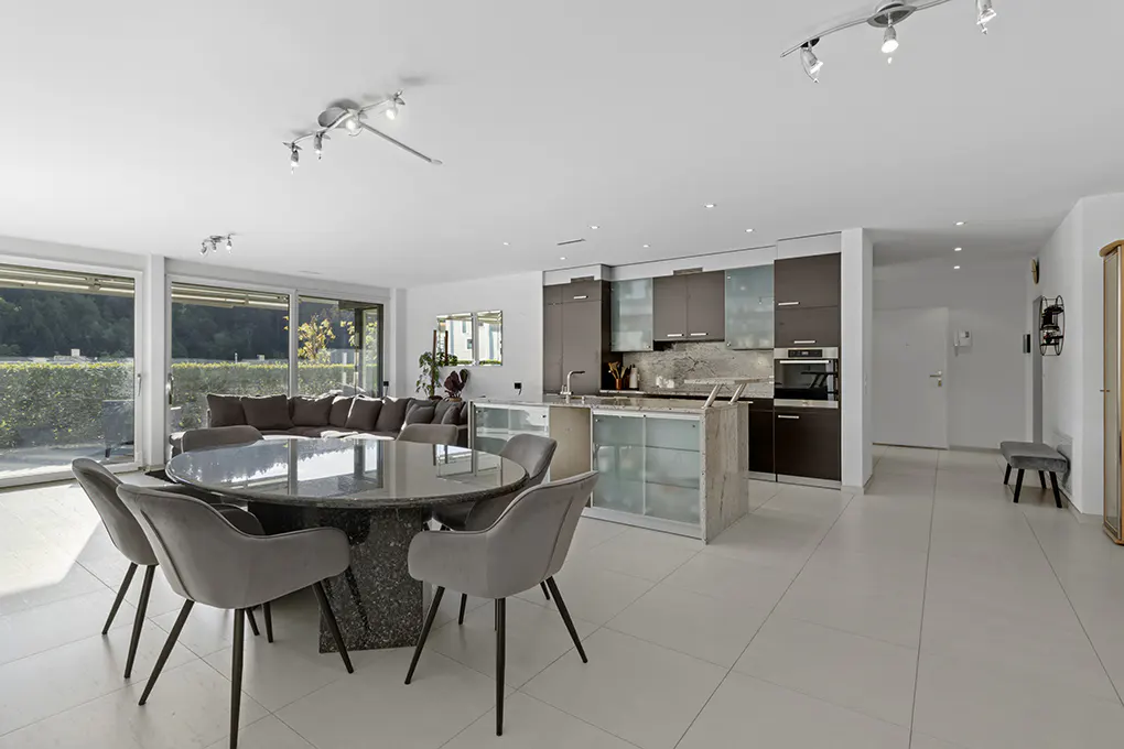 Open-concept living space with a round table, gray chairs, kitchen island, and a large window overlooking greenery.