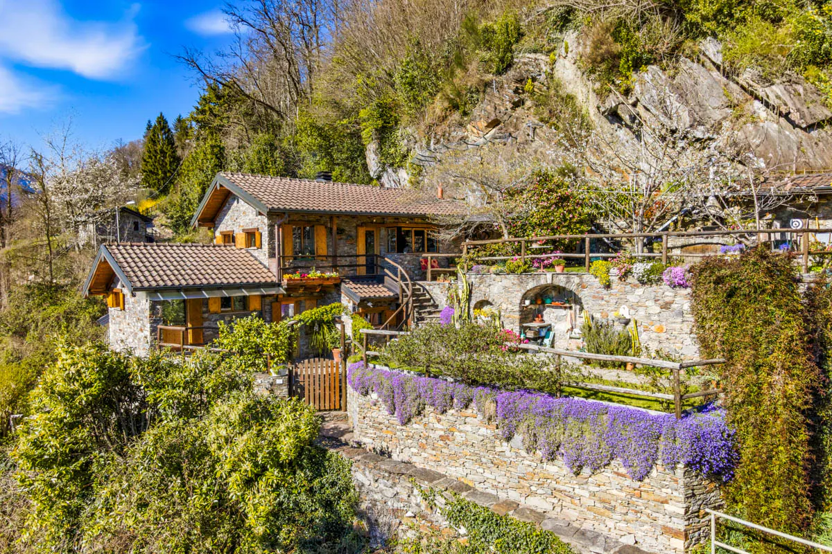 Stone house with brown shutters on a hillside, surrounded by lush greenery and purple flowers. Blue sky above.