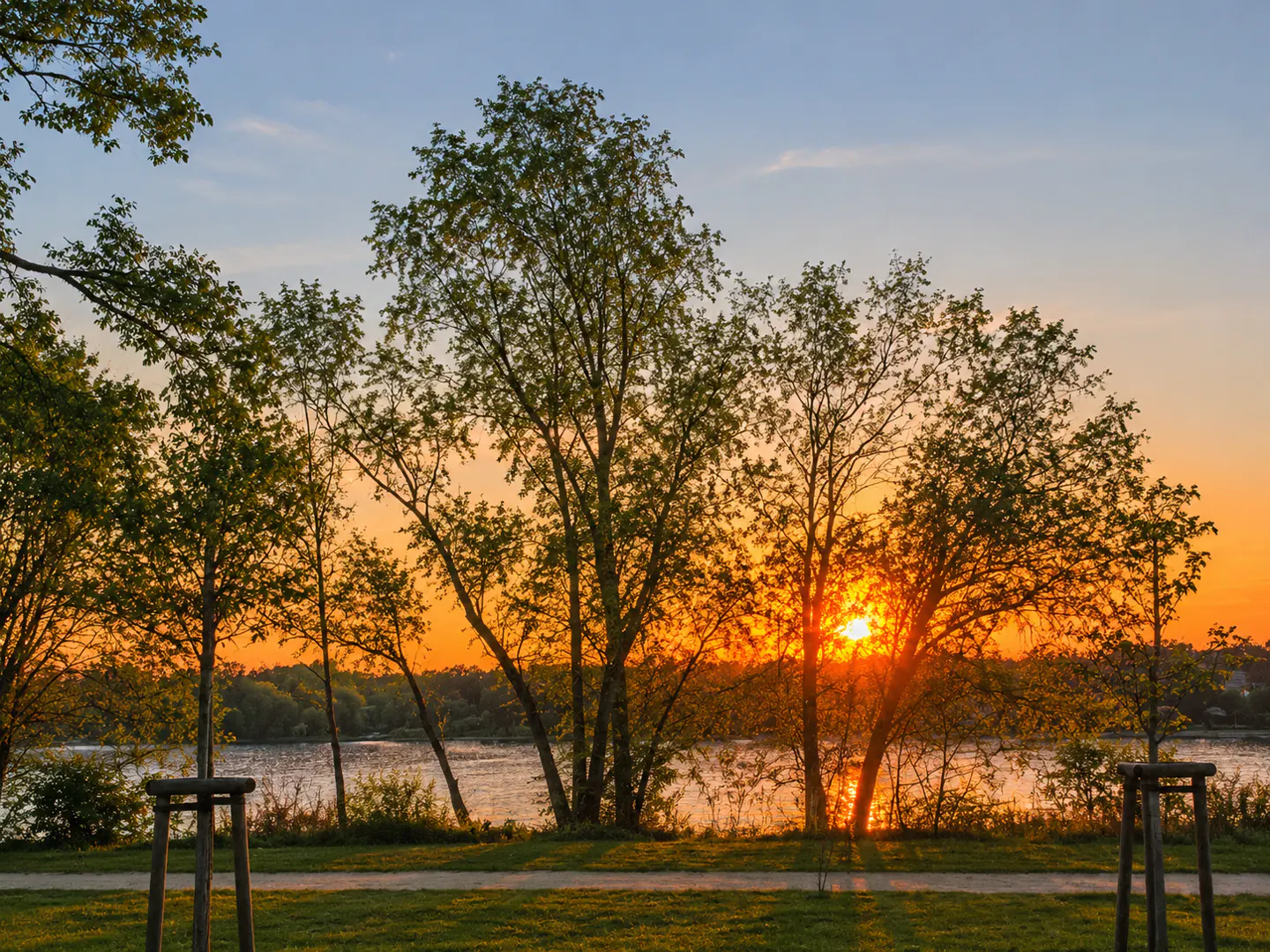 Sunset over a lake, viewed through trees. The sky is blue and orange. The sun reflects on the water.
