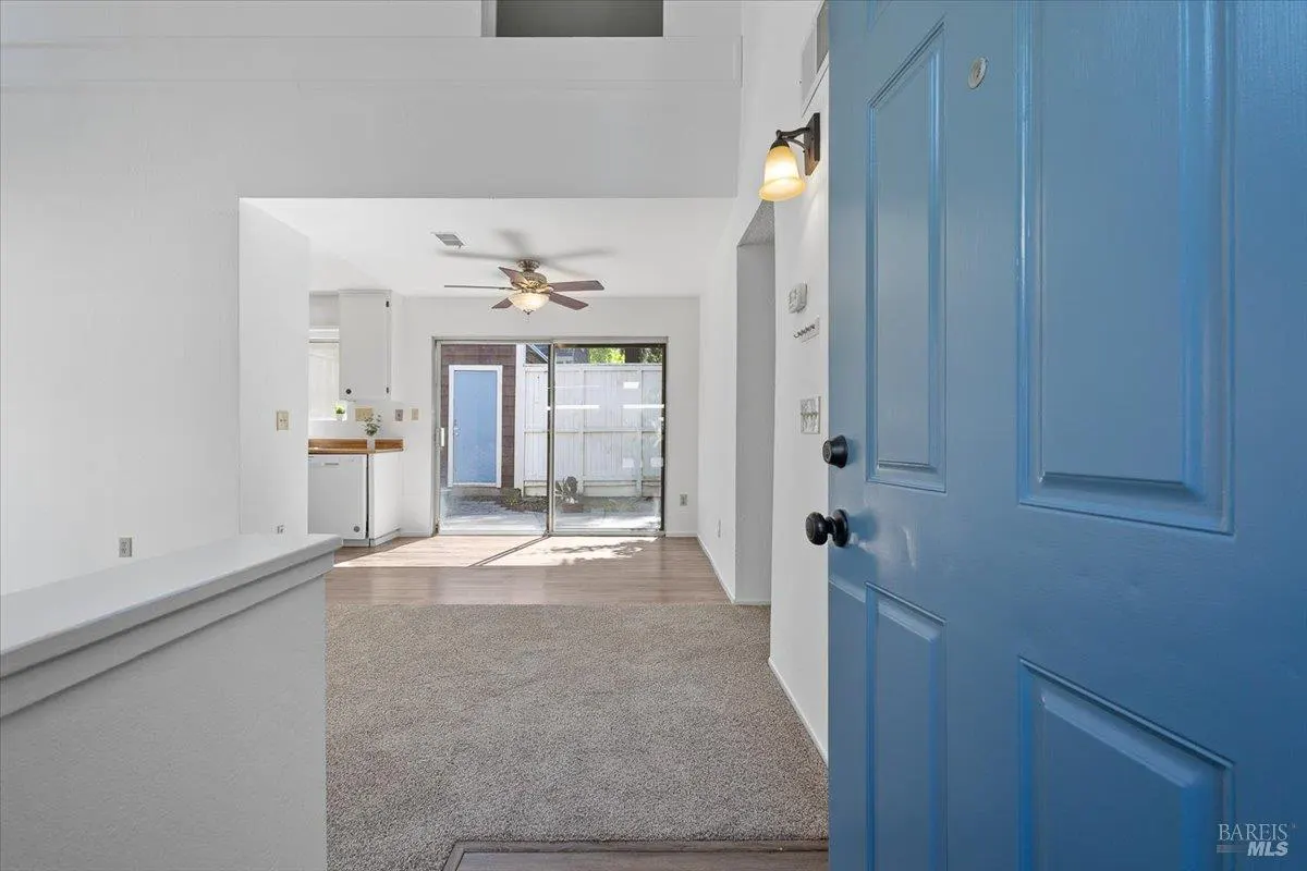 Open blue front door reveals a bright living room with beige carpet, white walls, and a sliding glass door to a patio.