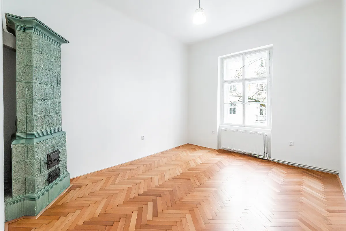 Bright, empty room with herringbone wood floors, white walls, and a green tiled fireplace. A window lets in natural light.