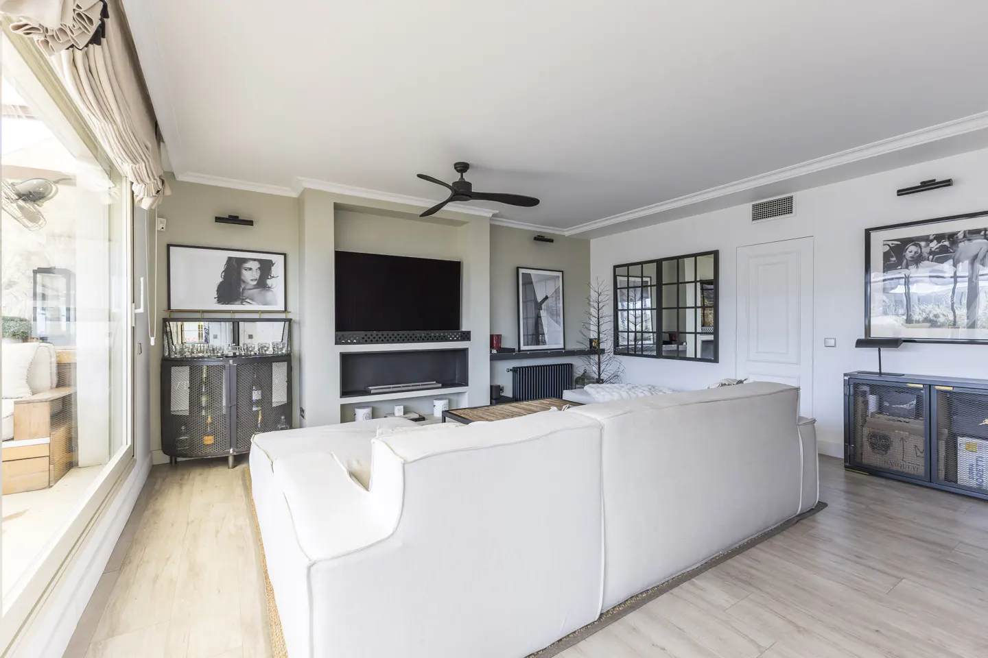 Bright living room with a white sofa, light wood floors, and a large TV mounted above a fireplace. Black and white photos decorate the walls.