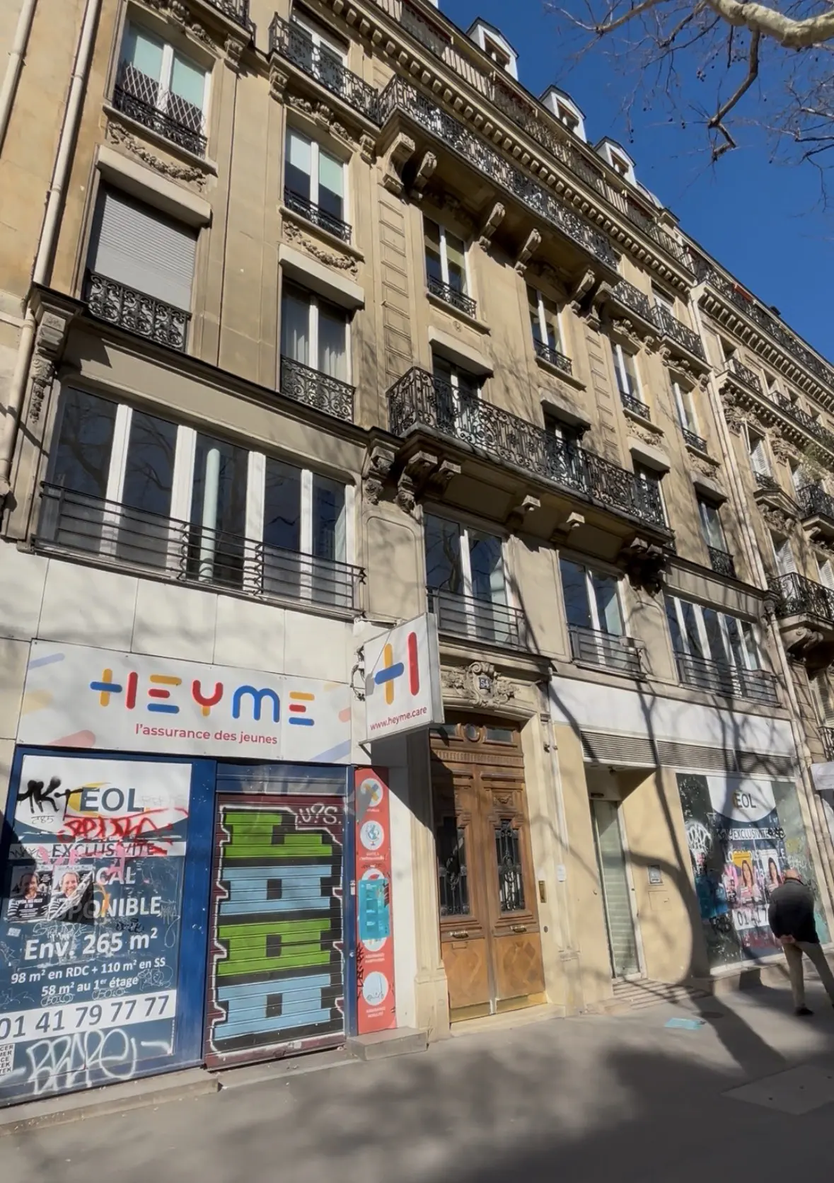 A tan, multi-story building with wrought iron balconies and a Heyme sign above a storefront with graffiti-covered posters.