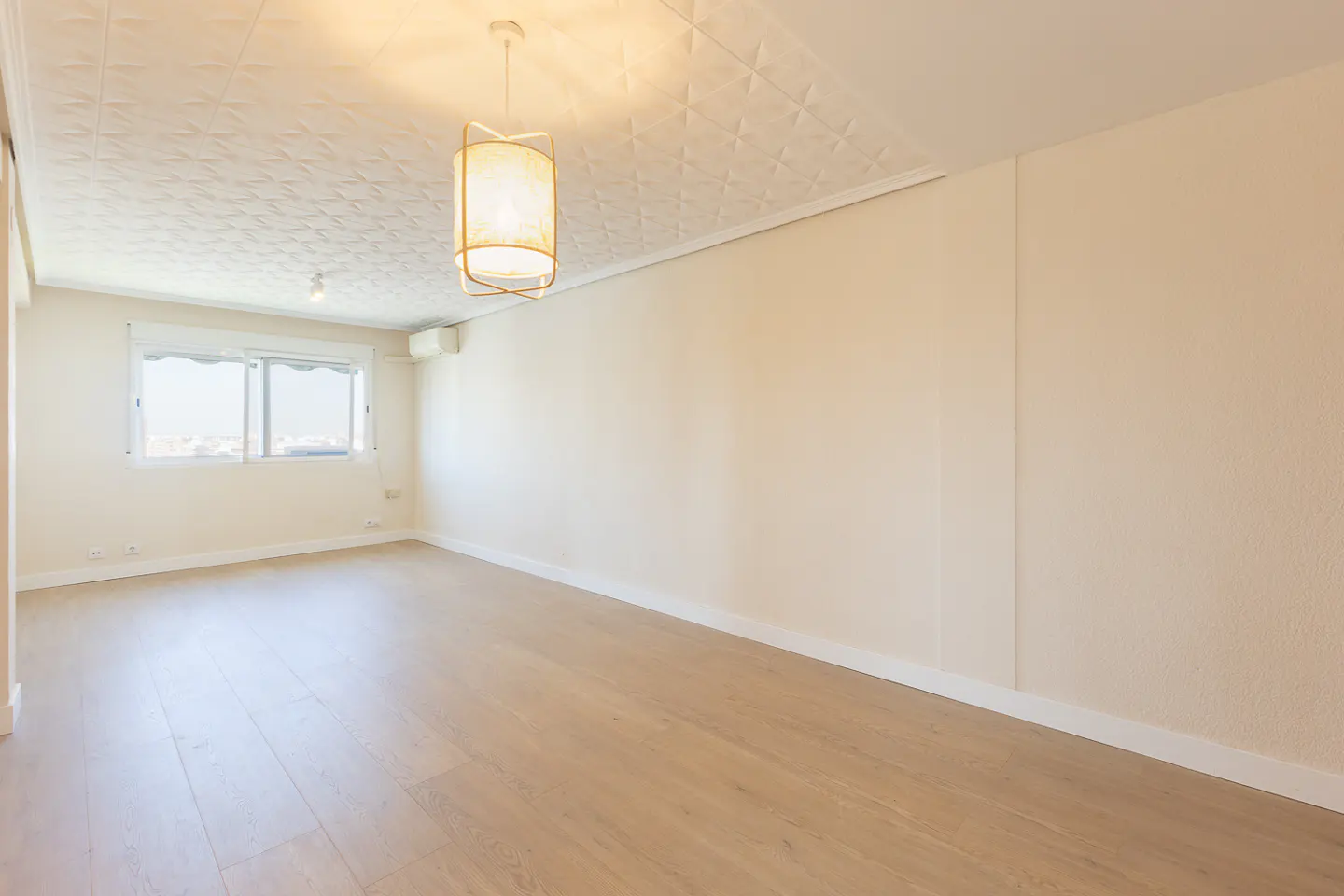 Bright, empty room with light wood floors, white walls, and a textured white ceiling. A window lets in natural light. A modern light fixture hangs from the ceiling.