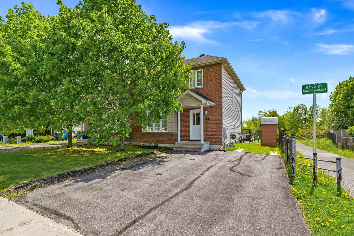Two-story brick and siding house with a paved driveway, large green tree, and a street sign under a bright blue sky.