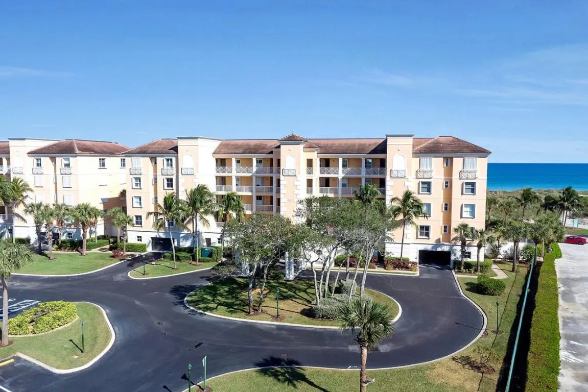 Aerial view of a peach-colored condo building with balconies, palm trees, and a circular driveway leading to the ocean.