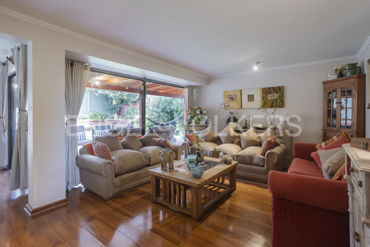 Living room with hardwood floors, two beige sofas, a red sofa, and a wooden coffee table. Large windows overlook a green garden.