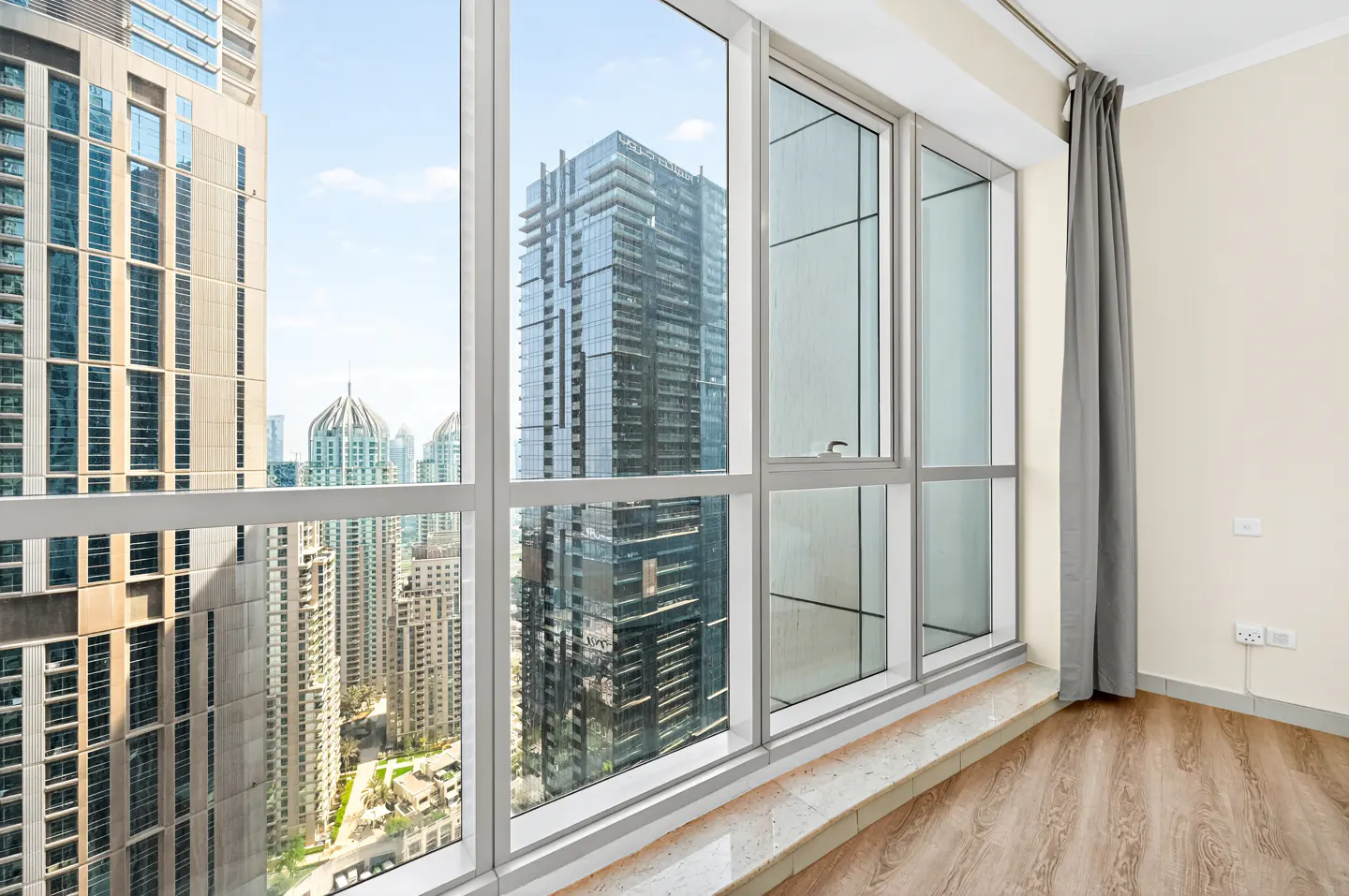 A bright room with a large window overlooking a city skyline. Gray curtains hang to the side of the window. Wood-look flooring.