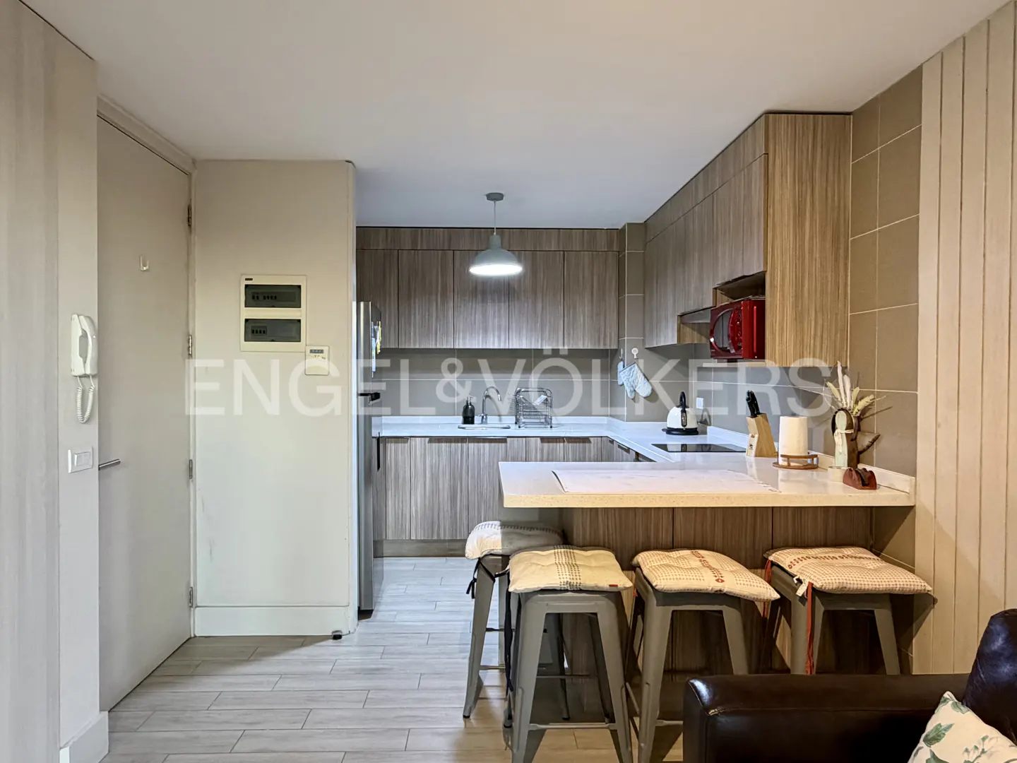 A modern kitchen with wood cabinets, a white countertop, and three stools at the breakfast bar. The floor is light gray tile.