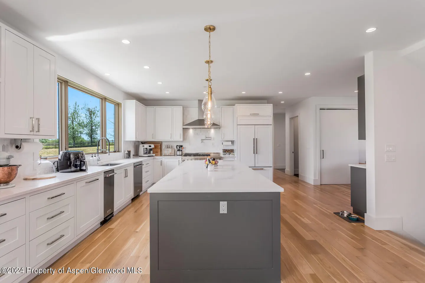 Bright, modern kitchen with white cabinets, gray island, and wood floors. A large window overlooks a green field. Gold pendant lights hang above the island.