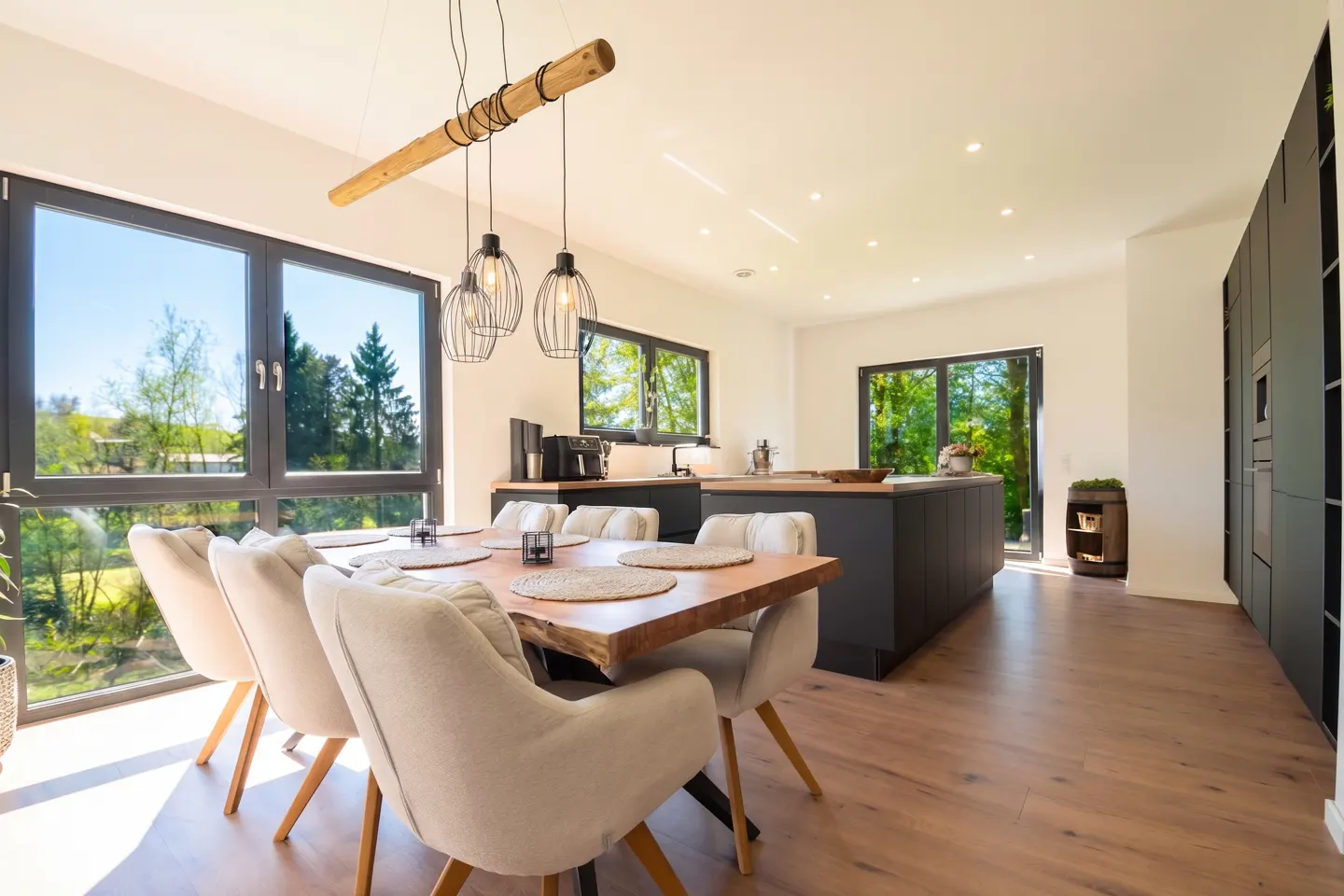 Bright, modern kitchen with a wood table, beige chairs, and black cabinets. Large windows offer a view of green trees. Unique light fixture above the table.