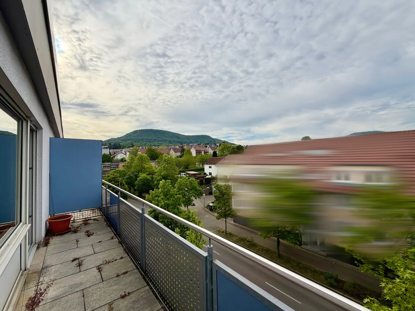 Balcony view with gray stone floor, metal railing, and blue panel. Distant green hill and cloudy sky. Street and blurred building visible.