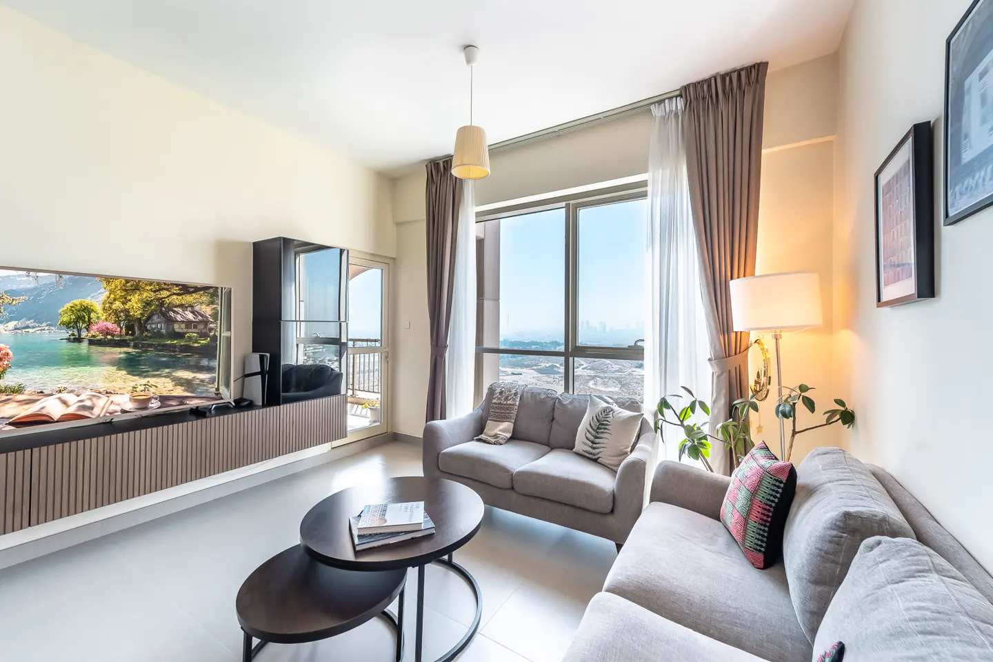 Living room with gray sofas, a TV showing a lake scene, and a large window with city views. A round coffee table sits in the center.